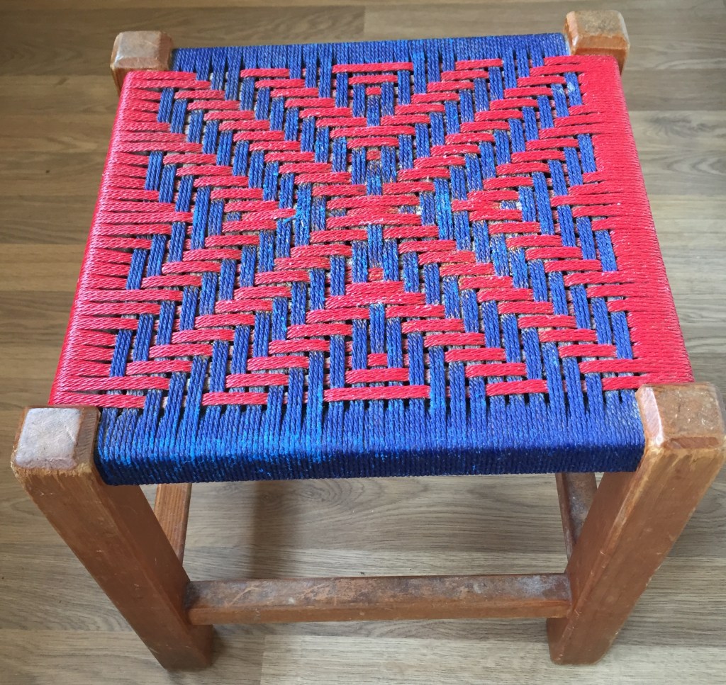 Small, square, flat stool with wooden legs. Seat has alternating blue and red pattern. From each edge, triangular arrow shapes of alternating colour and increasing size work towards the centre, which is a red X shape joining all the corners.