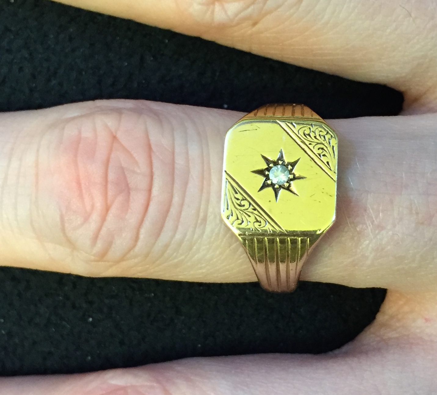 Signet ring. The top is square with rounded corners. The top-right and bottom-left corners have an ornate design. The centre of the ring has an 8-point star etched into it, with a diamond embedded in the middle.
