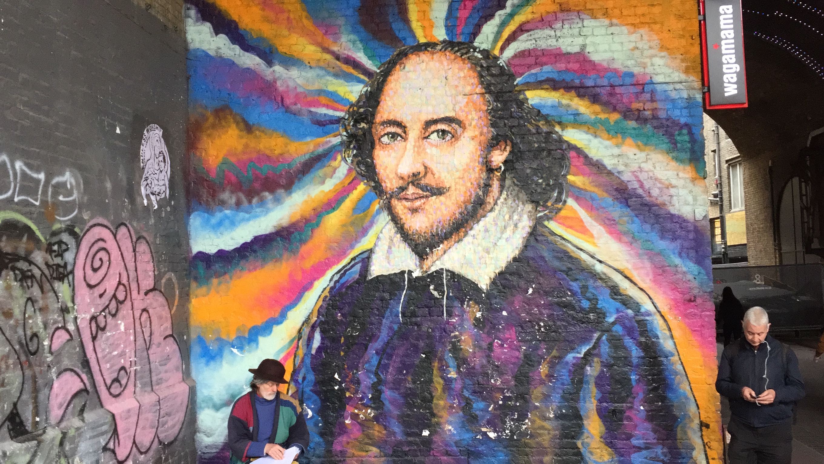 A large wall-sized painting of William Shakespeare, with a colourful swirly background. The artist is next to it at the bottom, still working on it.