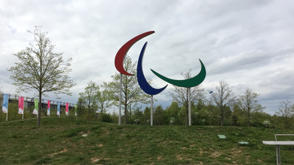 Large sculpture of the Paralympics symbol, made of 3 agitos marks in red, blue and green. It's held up on 3 poles, in the middle of the grass with trees in the background.