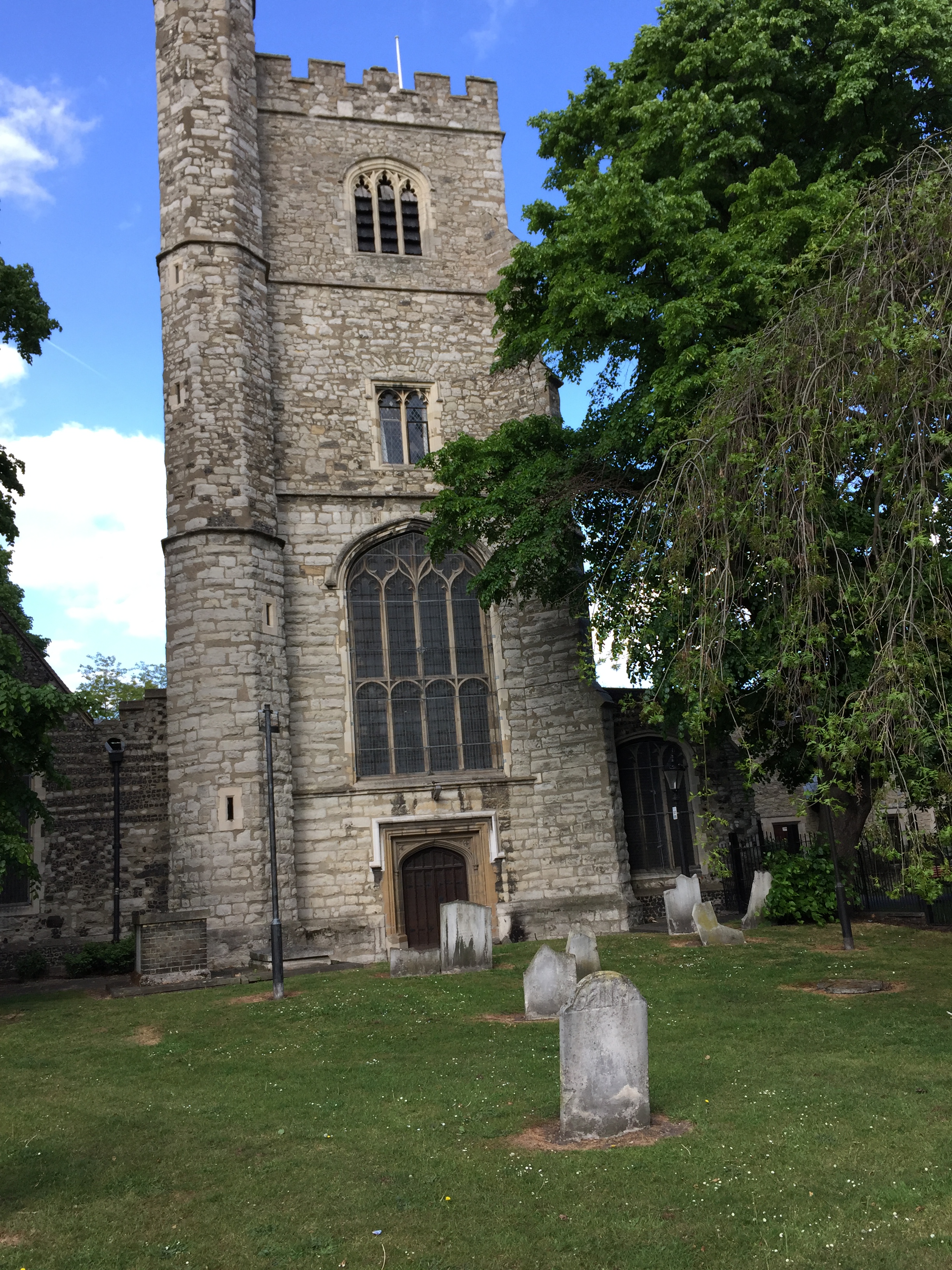 A large stone tower, called a curfew tower, at Barking Abbey. There is a doorway at the bottom, a very tall arched window above it, and much smaller windows on the two floors above that. A few gravestones are on the grass in front of the tower.