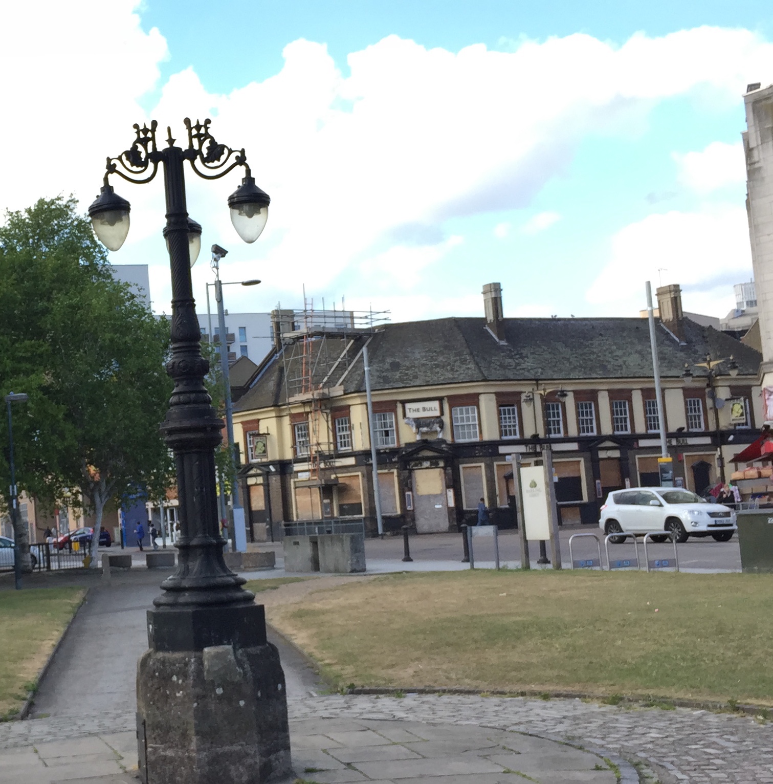 An ornate lamppost, with 3 lights hanging off it, with a pub called The Bull across the road behind it.