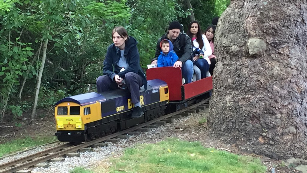 Adults and children sitting on a small model train as it moves along a track in Barking Park. The front engine is yellow and black, while the open carts being pulled behind it are red, with a few people squeezed tightly into each one.