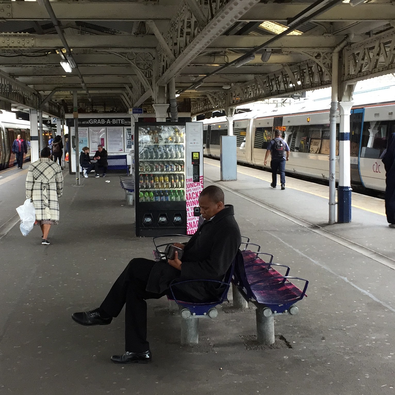 Two platforms at Barking Station, with a train waiting at each one. There is a wide concourse between the platforms, with a drinks vending machine and people sitting on benches.