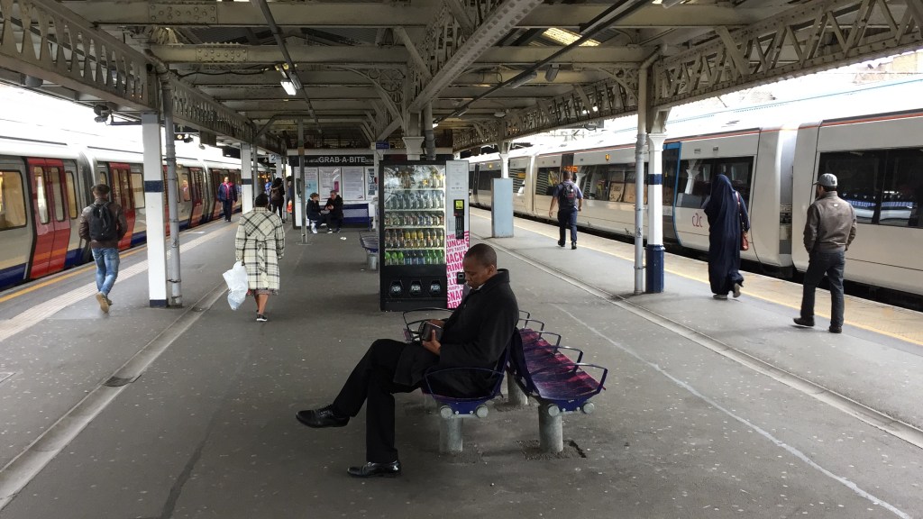 Two platforms at Barking Station, with a train waiting at each one. There is a wide concourse between the platforms, with a drinks vending machine and people sitting on benches.