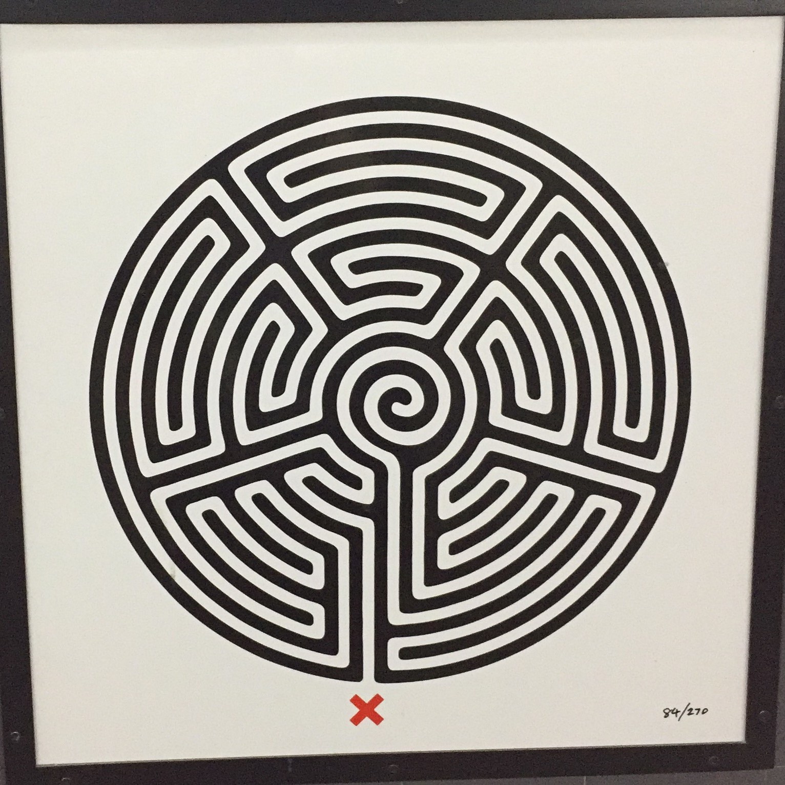 The labyrinth roundel artwork at Barking Station. This is a circular maze, in black with a complex white pathway winding through it, and a red X marking the starting point. Numbering in the corner of the sign indicates that this is number 84 of 270 such roundels, as there is one to be found somewhere at every station on the Tube network.