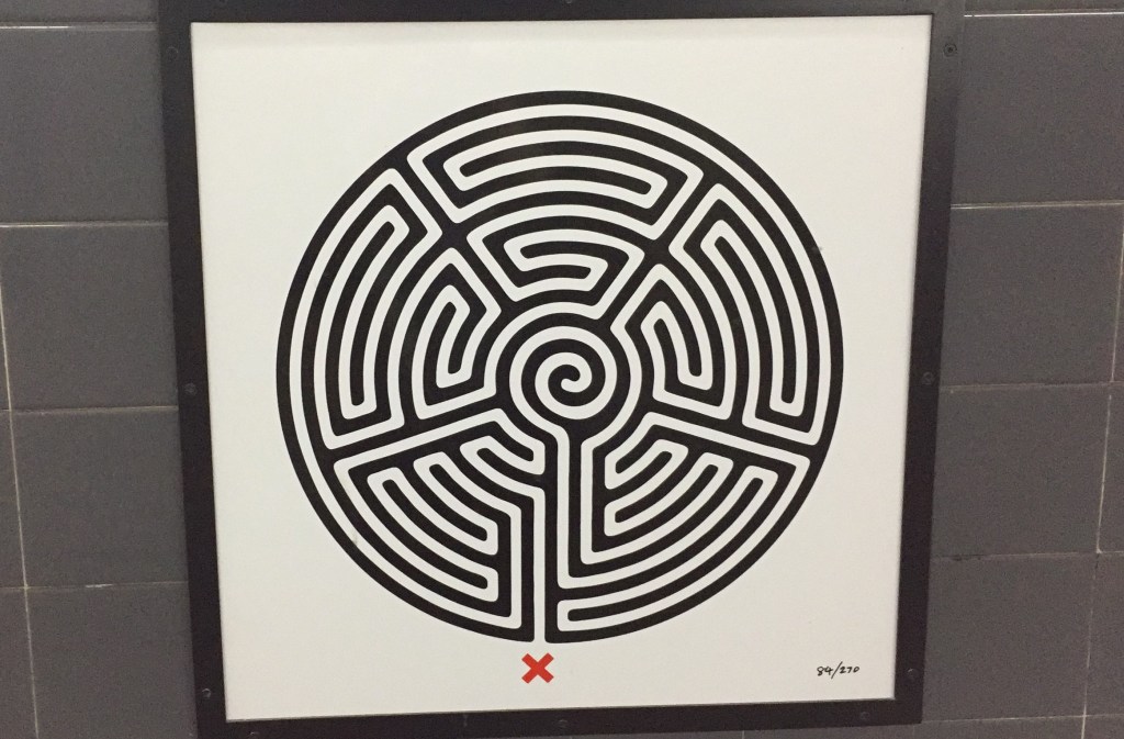 The labyrinth roundel artwork at Barking Station. This is a circular maze, in black with a complex white pathway winding through it, and a red X marking the starting point. Numbering in the corner of the sign indicates that this is number 84 of 270 such roundels, as there is one to be found somewhere at every station on the Tube network.