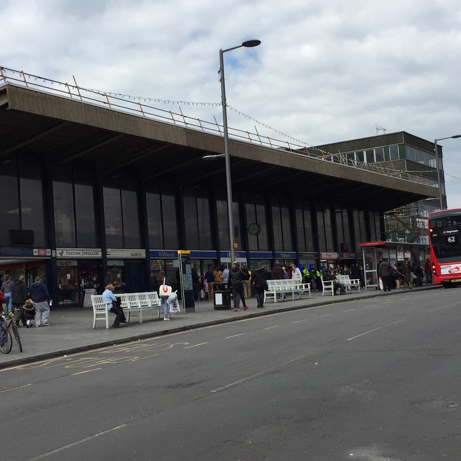 Full view of the exterior of Barking Station from across the street. It is a long building, with the station entrance and shops on the lower level, tall windows above them and a rectangular canopy jutting out from the roof along the entire length.