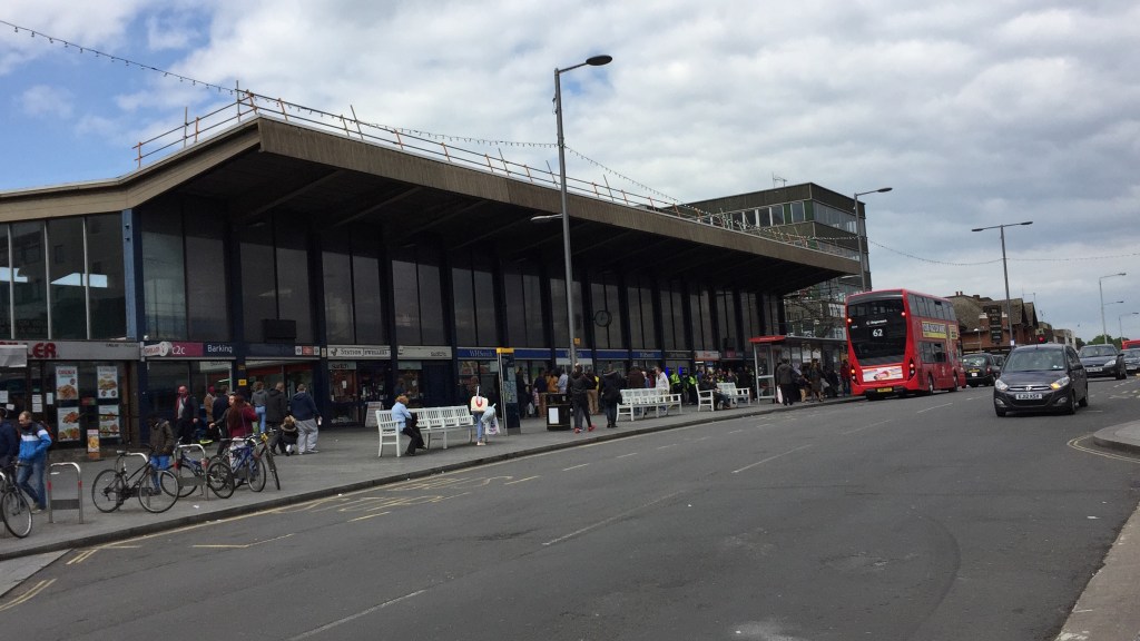 Full view of the exterior of Barking Station from across the street. It is a long building, with the station entrance and shops on the lower level, tall windows above them and a rectangular canopy jutting out from the roof along the entire length.
