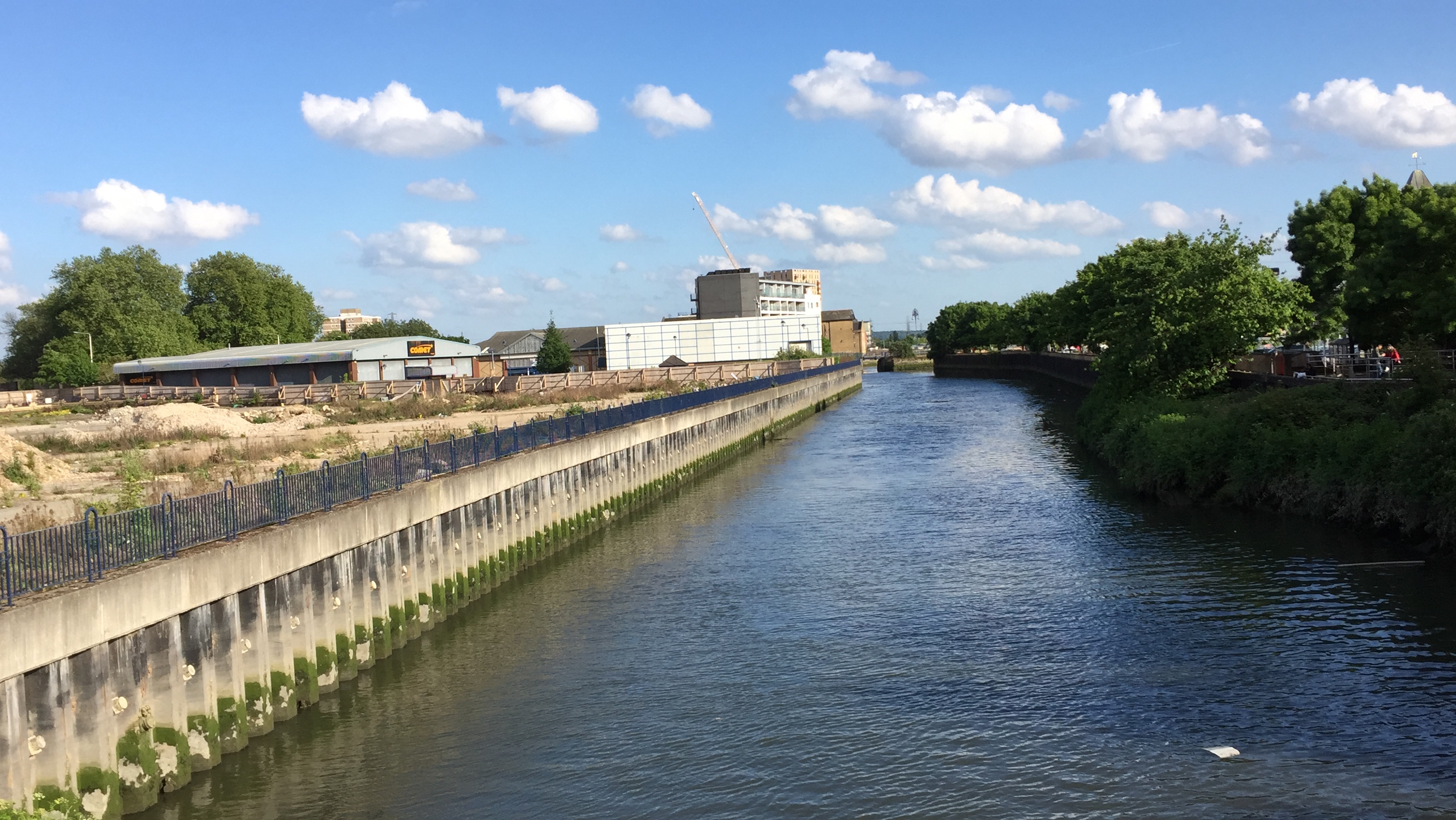 View along the River Roding in Barking, with trees on the right hand side, and a few buildings including a Comet warehouse on the left side.