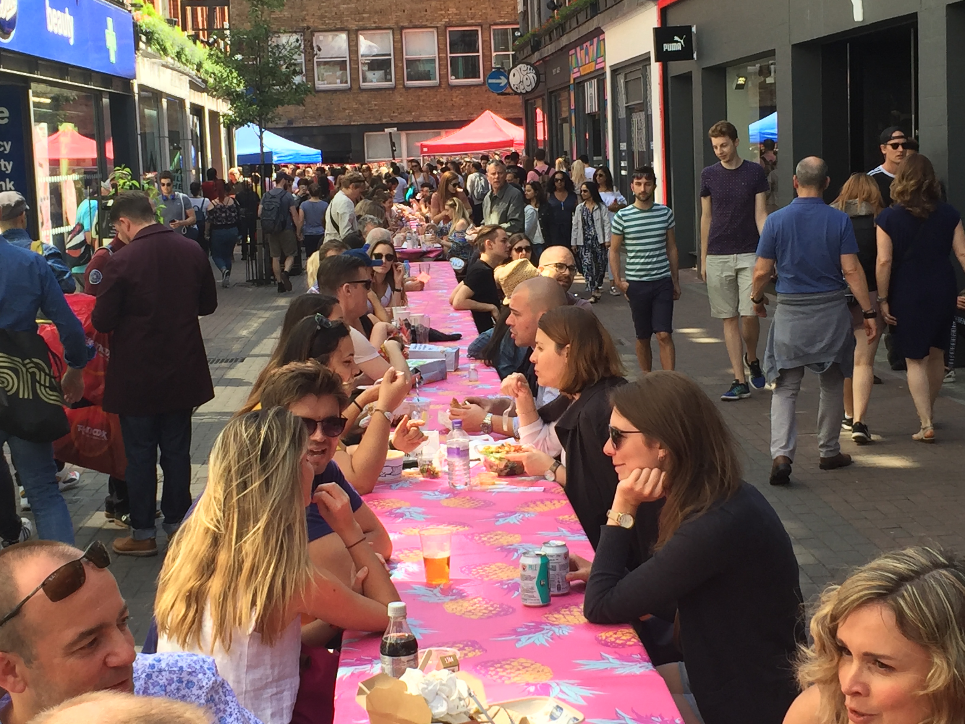 People sitting, chatting and eating at long narrow picnic tables covered in pink tablecloths in Carnaby Street.