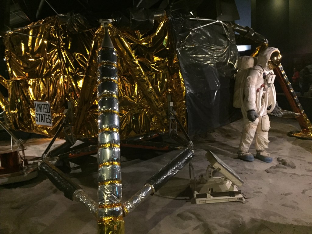 An astronaut standing in front of a large moon lander craft, the body of the craft covered in reflective gold foil. A white sign with black text attached the craft says United States.