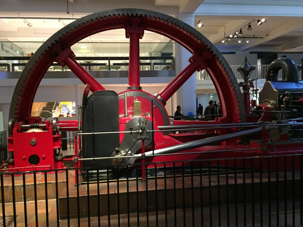 A very large red wheel attached to a steam engine in the Science Museum.