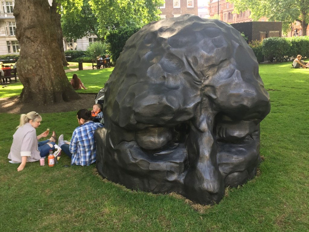 A family sit on the grass next to a giant bronze sculpture of a man's head in Cavendish Square. The head is only visible from the nose upwards, as if the rest of the face is buried below the ground. The sculpture is called Visitor by David Breuer-Weil.