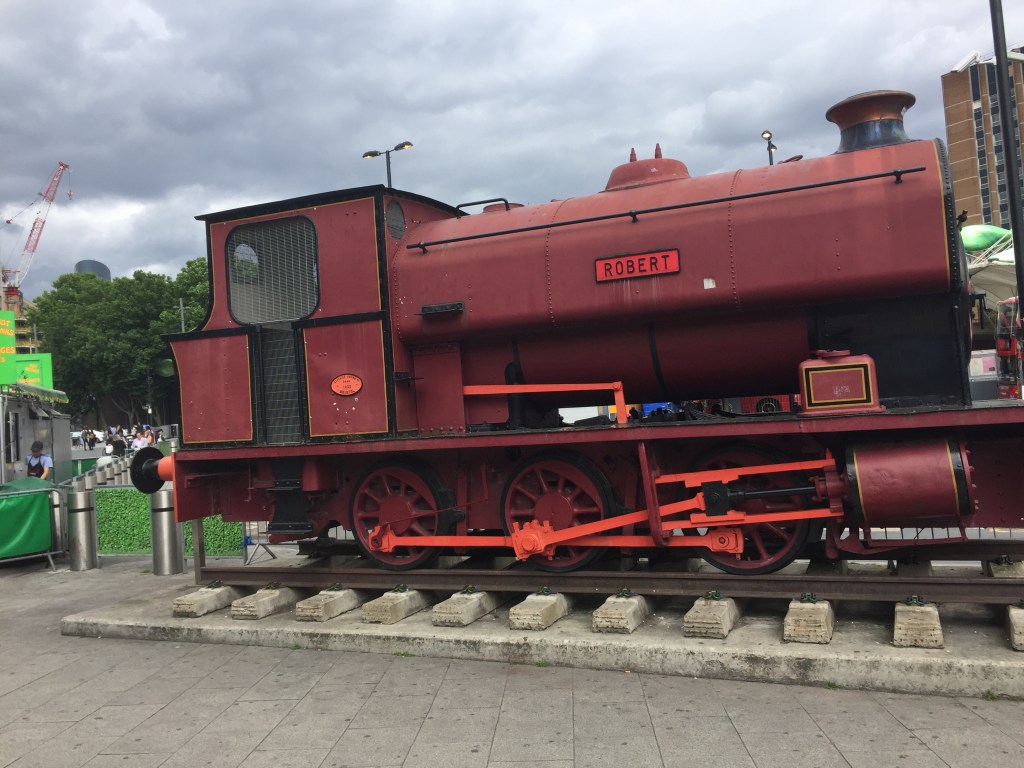 A large red steam train, with the name Robert in a small plaque on the side, sits on a small section of railway track placed outside Stratford Station.
