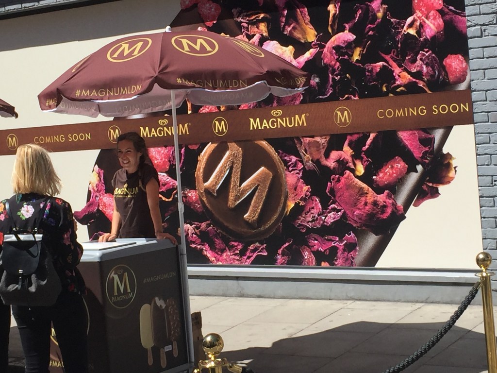 Sheltering from the sun under a Magnum themed umbrella, a lady at the Magnum stall serves a customer. Behind her, the shop wall is filled with an image of a fruit flavoured Magnum. A horizontal banner across the image has the Magnum logo and the words Coming Soon.