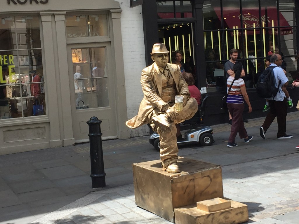Completely covered in gold, a man dressed in a jacket, shirt, trousers and hat holds a beer while in a seated position in mid-air. His right foot is the only part of him in visible contact with the box below, as he's resting his left foot on his right knee.