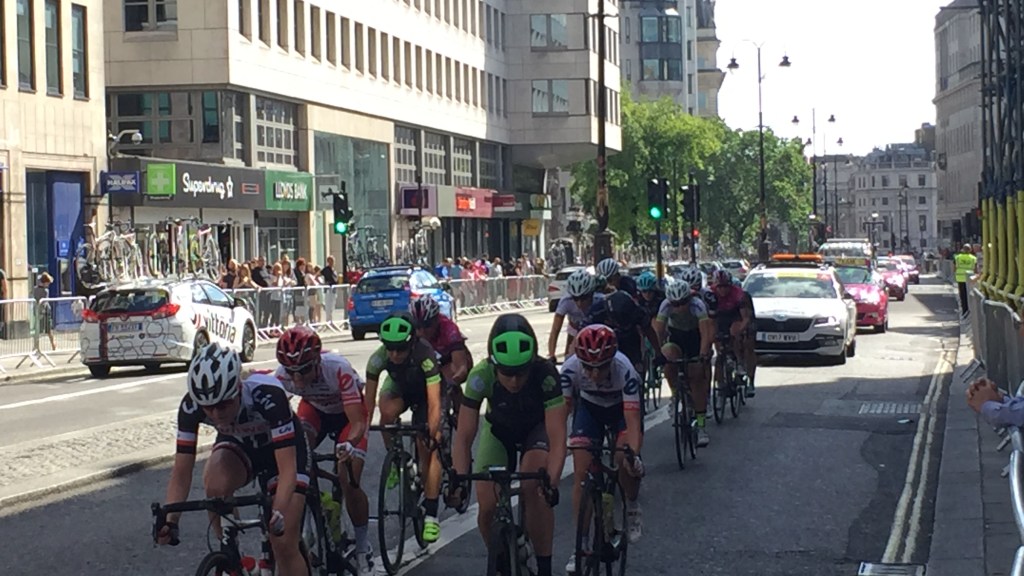 As part of the OVO Energy Women's Tour Stage 5, a group of cyclists race down the wide Strand road in London, with cars following behind. A small crowd is gathered behind the metal railings that stretch along the edge of the pavement.