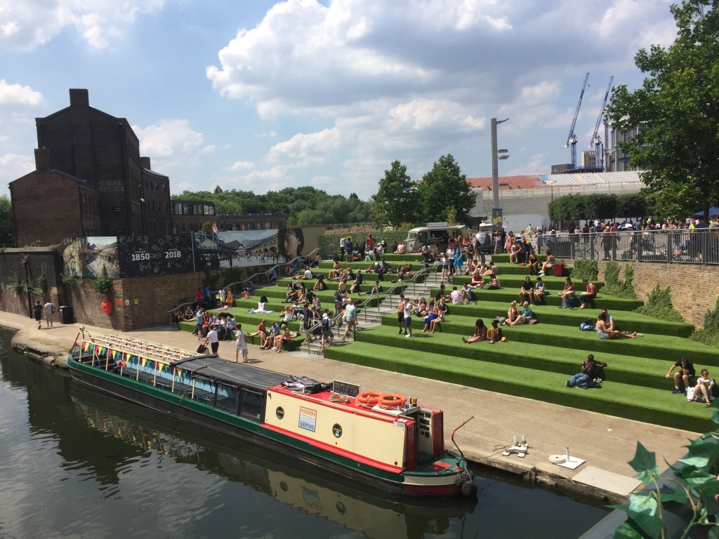 Kings Cross Canal with a long barge parked by the path. Next to this, crowds of people are sitting on on a very wide set of big green steps, with food and drink stalls behind them at the top.