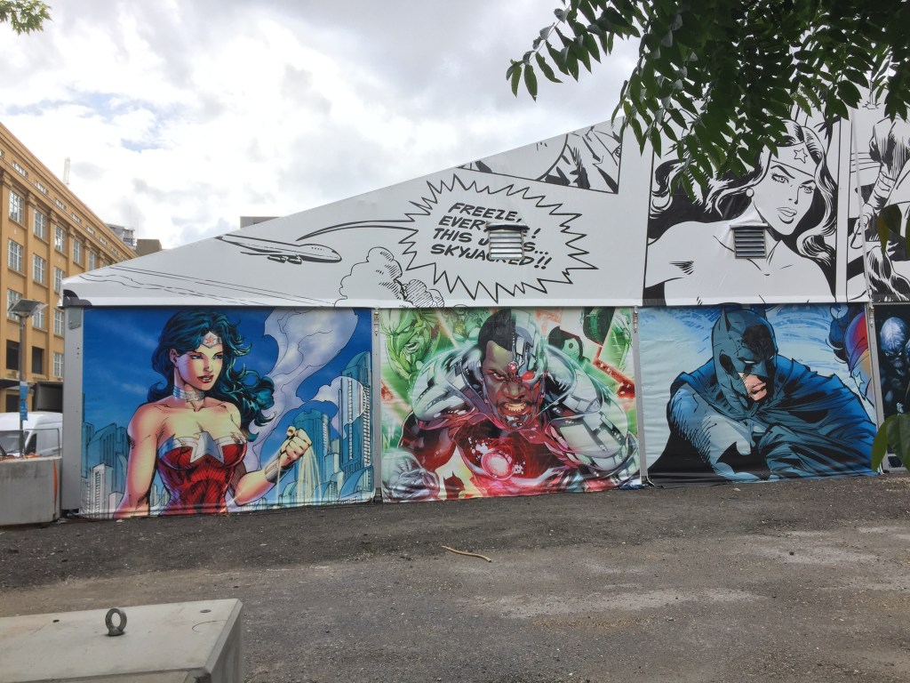 A mural in the form of a comic strip, stretching along the side of a single-floor building. Along the wall are colourful pictures of various superheroes, such as Batman. On the white roof above, a speech bubble comes out of a plane saying Freeze everyone! This jet is skyjacked!", with black & white images of characters to its right.