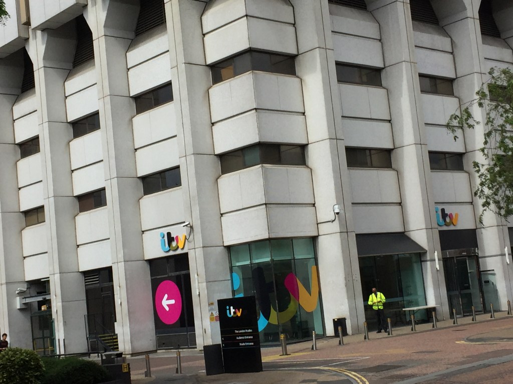 Bottom corner of the ITV studios tower, with the ITV logo splashed across the lowest corner window. A large pink circle with a white arrow points left on one of the lower left windows, while a tall black sign next to this points to the audience and studio entrances on the left of the building. A security guard is also standing outside in a hi-vis jacket.