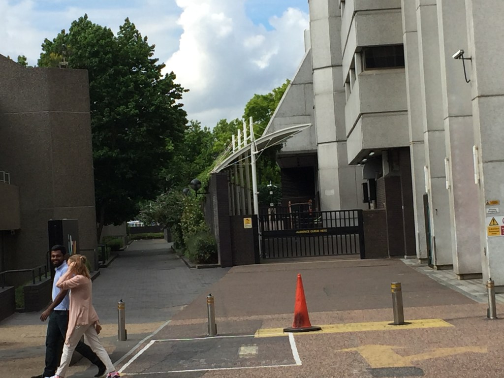 Black barred gate with the words Audience Queue Here on the left side of the ITV Studios tower when viewed from the road. On the other side is a caonpy stretching out from the building, under which audiences members walk to get inside.