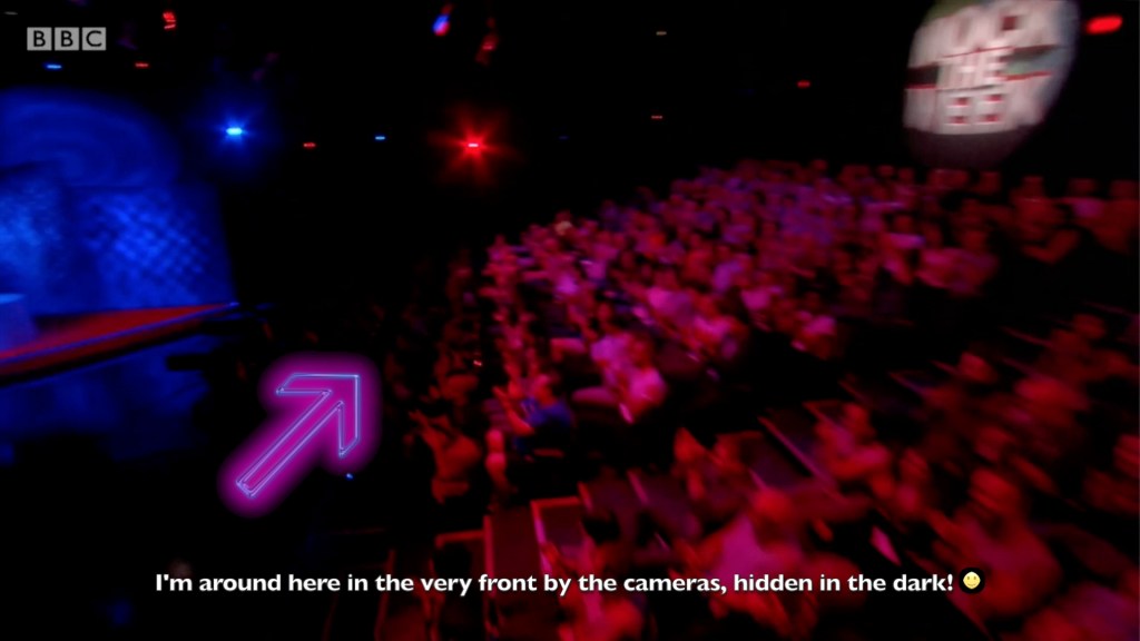 Screenshot of the audience from Mock The Week. An arrow points to the very front row, on the right of the central steps when facing away from them. The text reads - I'm around here in the very front by the cameras, hidden in the dark!