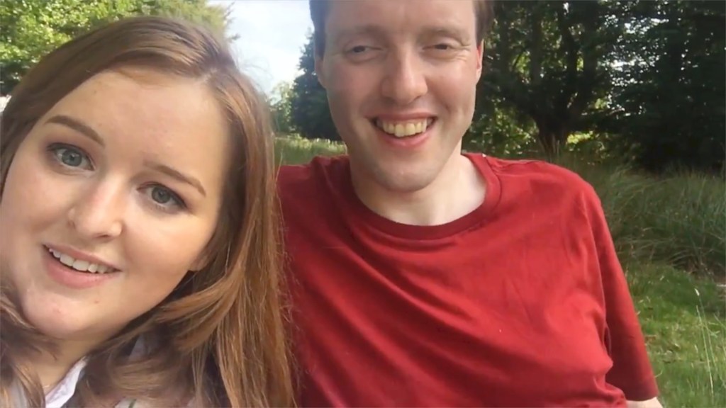 Selfie of Glen with Emily Davison, sitting together on some grass in the shade, with trees and sunshine in the background.