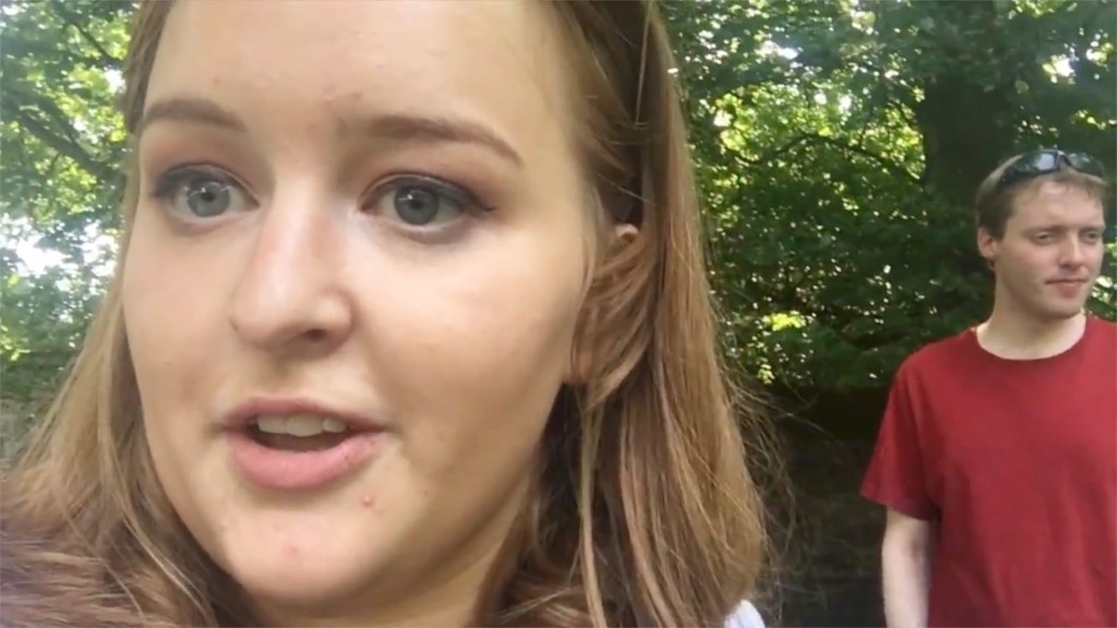 Emily talking to the camera, with Glen walking just behind her, in the shade under some trees.