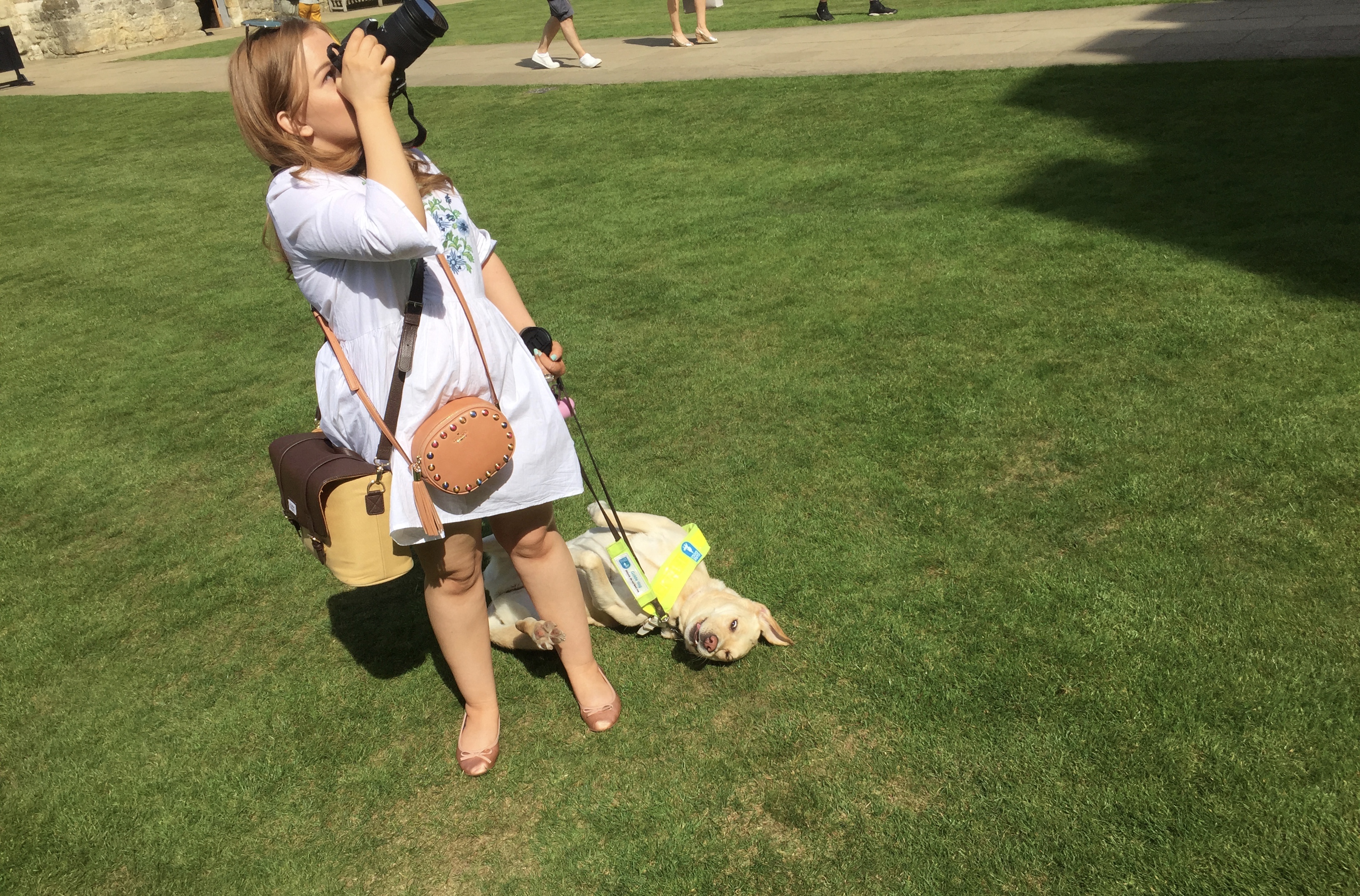 Emily looking upwards as she takes a photograph, with her guide dog Unity looking happy as she rolls around on the grass beside her.