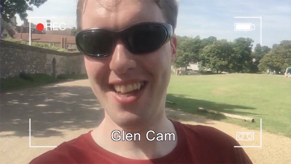 Glen wearing sunglasses and smiling at the camera in the sunshine. Overlaid on the image is a mockup of a camera recording screen, with 4 right-angled corner marks, a recording symbol top-left, a battery level indicator top-right, and the words Glen Cam centred at the bottom.