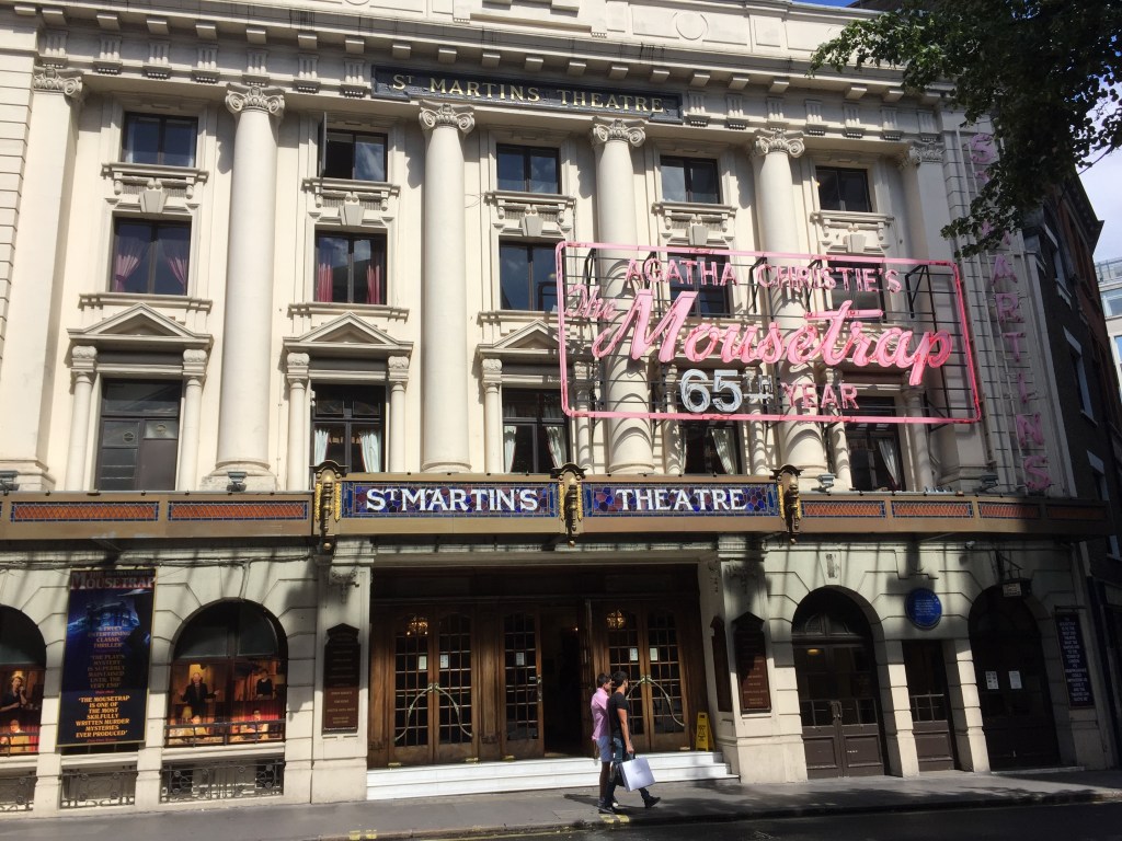 The outside of St Martin's Theatre. The theatre name is in white text on a blue background across the top of the doorway. Above this, hanging in front of the windows, is a large sign that says Agatha Christie's The Mousetrap, 65th Year. The play's title is in pink script lettering, the rest is in pink block capitals.
