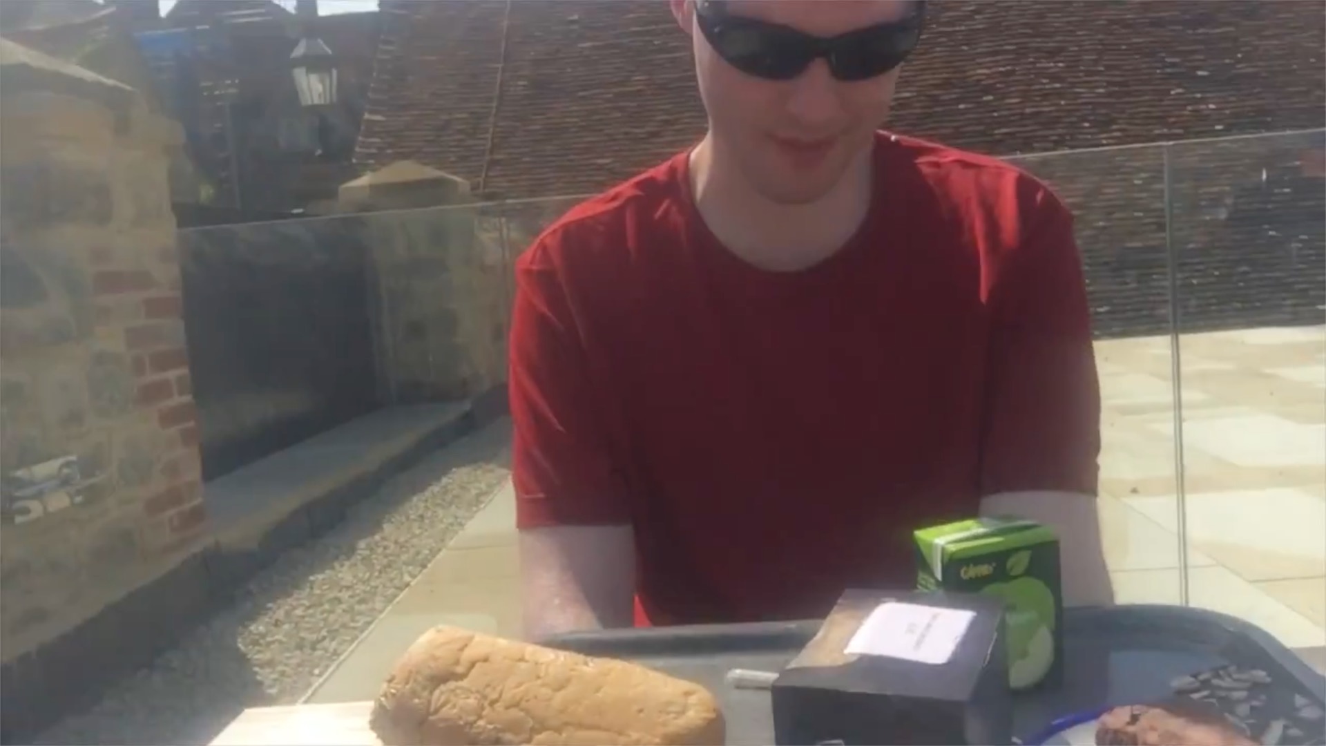 Me sitting at a table outside in the sunshine, with a tray of lunch in front of me, including a large roll, a sandwich, two cakes, and a carton of apple juice.