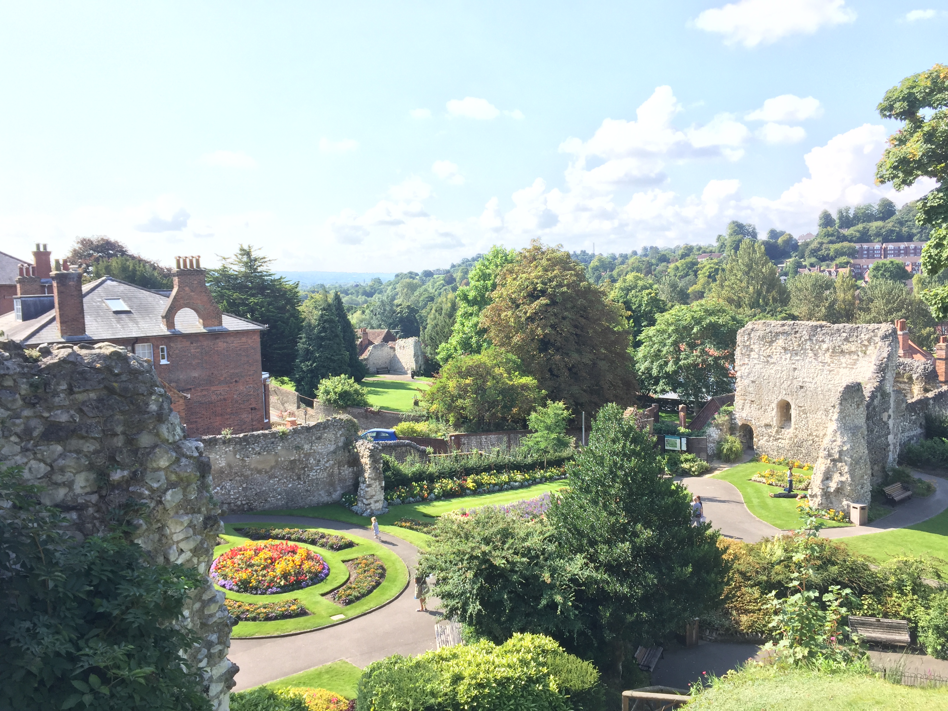 View from the roof of Guildford castle, across a garden bathed in sunshine, featuring a round flowerbed, tall trees, and parts of the old walls surrounding the castle.