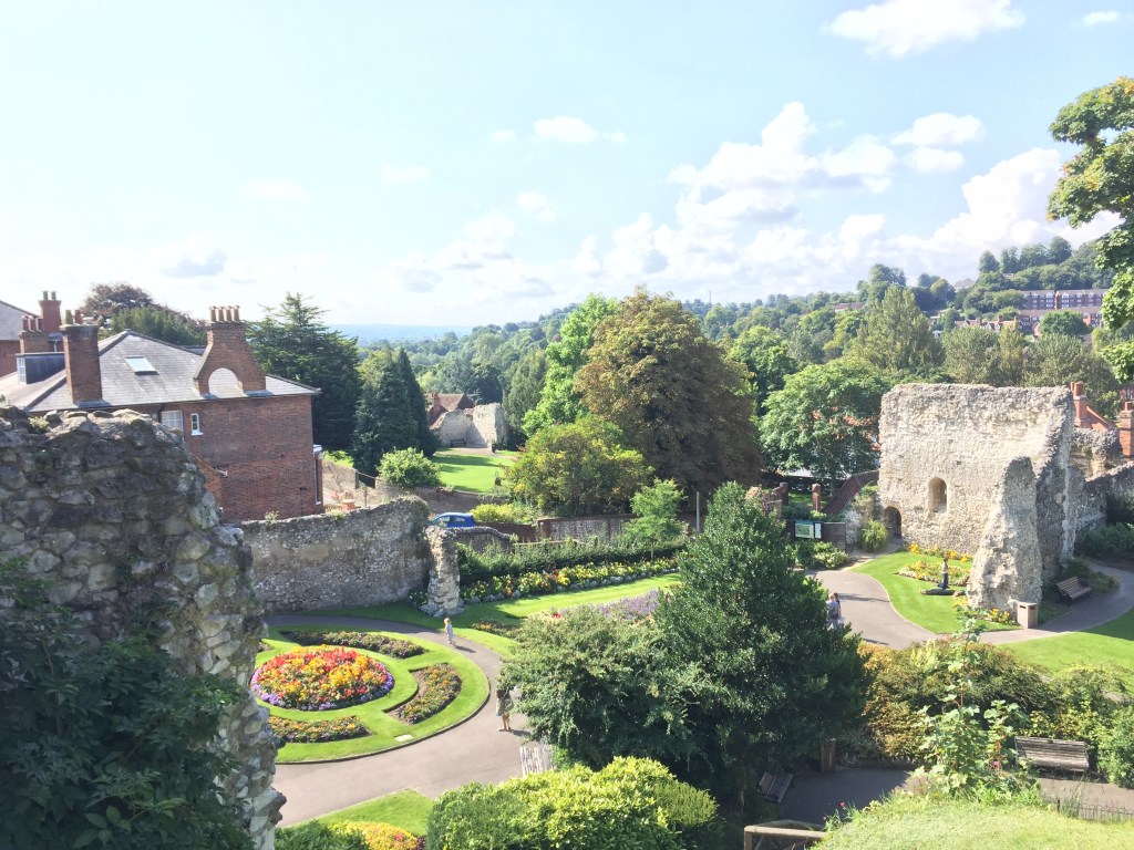 View from the roof of Guildford castle, across a garden bathed in sunshine, featuring a round flowerbed, tall trees, and parts of the old walls surrounding the castle.