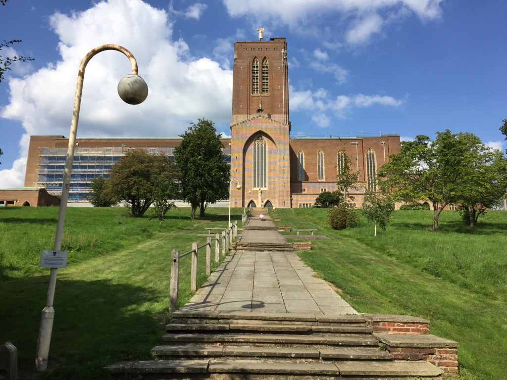 Guildford Cathedral - a very wide building with a very tall tower in the middle. The approach consists of groups of 5 steps, separated by long flat sections, gradually going up the hill, with grass and a few trees on each side.