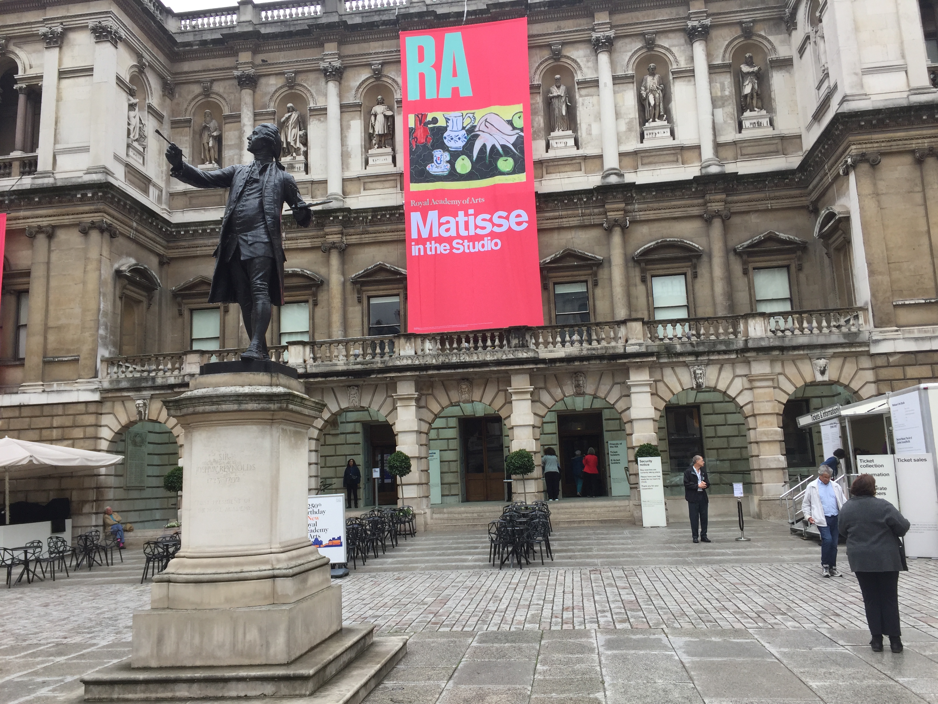 The Royal Academy of Arts building. A tall red banner hangs from the roof in front of it, with the wording Matisse In The Studio in large white letters and a picture of one of Matisse's collage artworks.