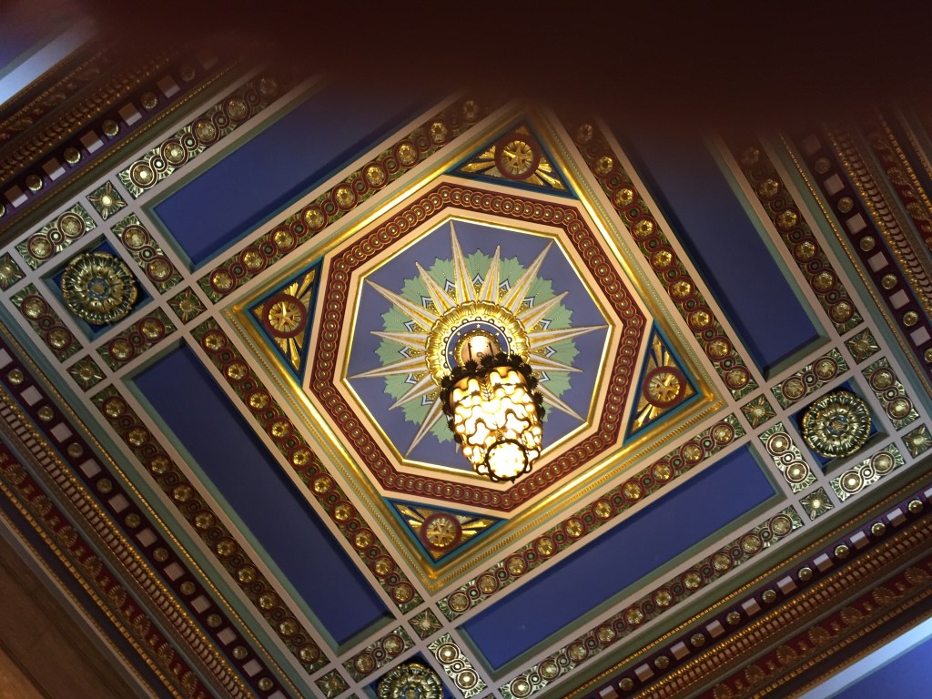 Ornate ceiling in Freemasons Hall. A decorative two-tiered light hangs beneath a large starburst motif on the ceiling, contained within an decorative octagon border, all of which is surrounded by a square pattern. Gold and blue are the dominant colours.