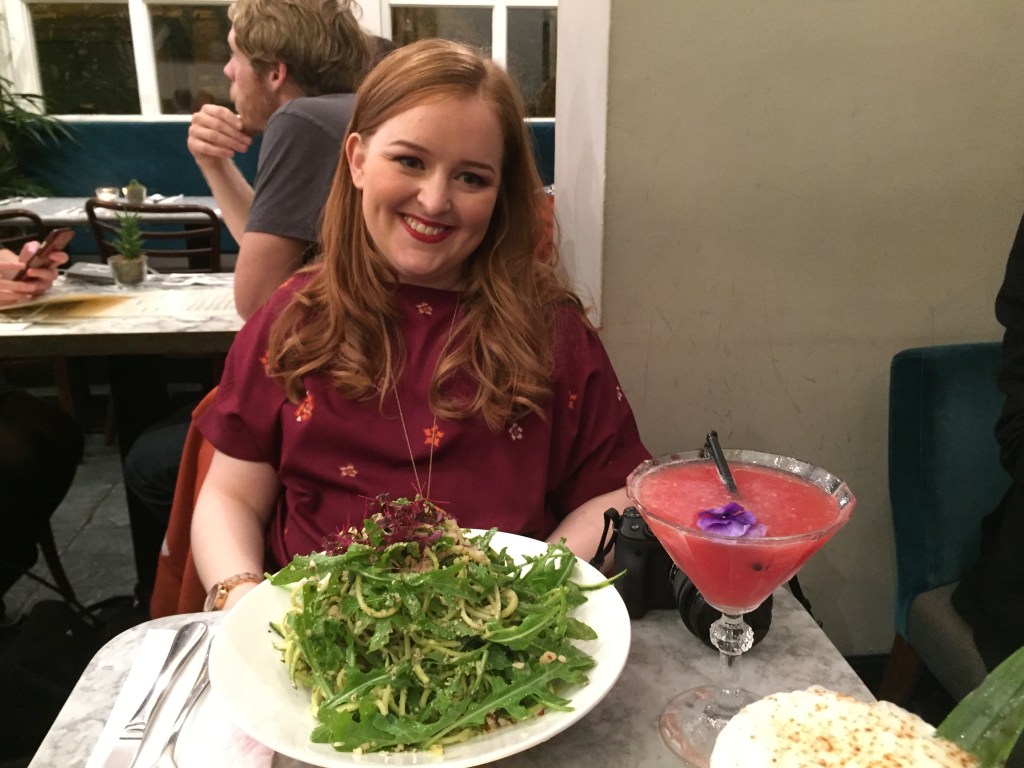 Emily smiling and wearing a burgundy coloured top with orange and pink flowers. On the table in front of her is a big plate of courgette pasta, and a cranberry cocktail.