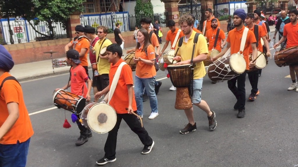 Group of people walking in a carnival, wearing orange t-shirts and playing drums.