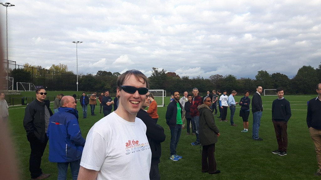 Me standing in the middle of a football field surrounded by many other people. I'm wearing sunglasses, and a white t-shirt with a colour logo saying All The Stations.
