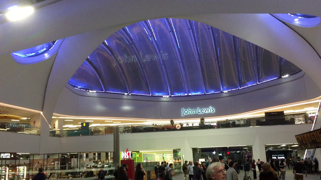 View from the ground floor of Birmingham New Street station. On a mezzanine level ahead is a John Lewis store. Above is a large curved ceiling with long, dark blue glass panes, through which can be seen another John Lewis sign.