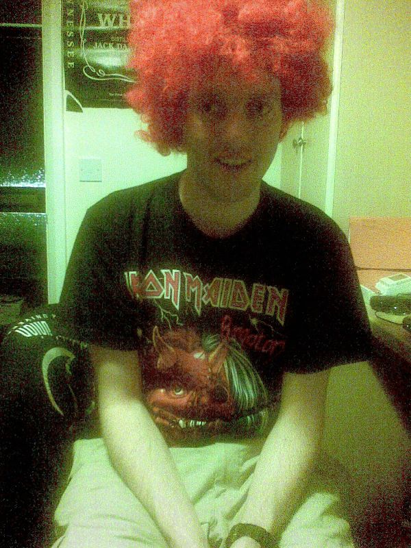 Me smiling while wearing a red wig and a black t-shirt featuring the logo and image of the band Iron Maiden.