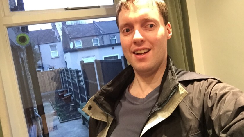 Selfie of me wearing a coat, standing in front of a window looking out at the paved back garden, with the backs of other houses beyond the wooden fencing.