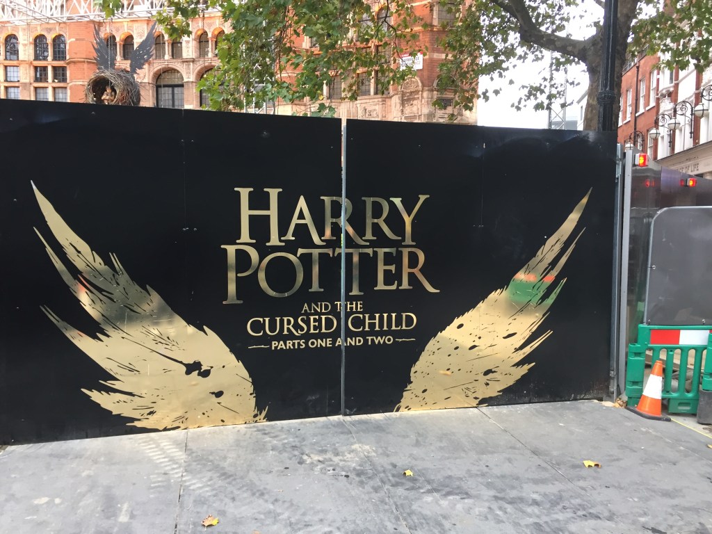 Large black hoarding with large gold lettering saying Harry Potter and the Cursed Child, Parts 1 and 2. This is framed by large gold wings rising from the bottom, one on each side of the text.