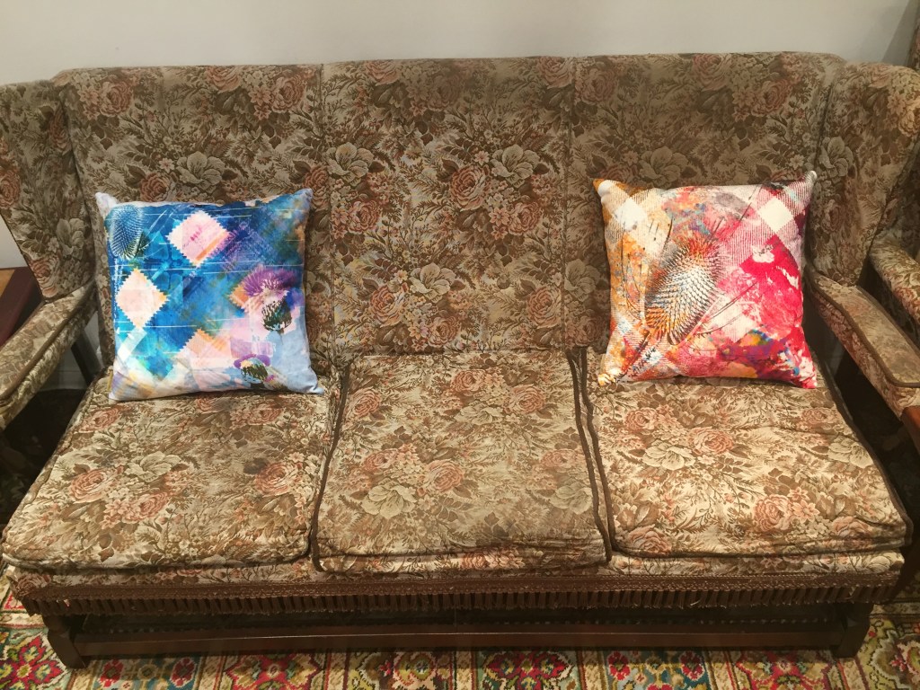 Two cushions at each end of a brown 3 seater sofa. Both cushions are very artistic, with splashes of various colours all over, and a black stencilled motif of a Scottish thistle. The left cushion is mainly blue with a large white diamond shape on each edge. The right cushion is a mixture of red and gold colour, with a couple of white diagonal lines in the background forming a cross in the centre.