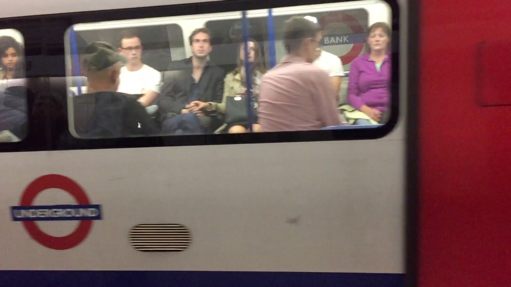 Tube train carriage viewed from the outside, with the Underground logo on the outside, and passengers seated on the inside. Through the back window of the carriage is the sign for Bank station.