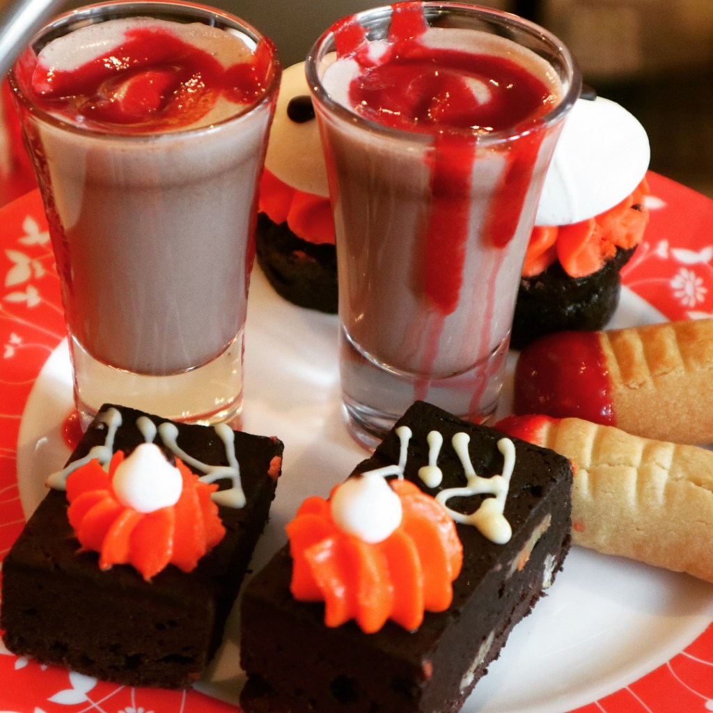 A plate containing small sweet treats in pairs, including 2 chocolate milkshakes containing raspberry coulis to look like blood, and 2 chocolate brownies iced with the letters R.I.P and topped with a pumpkin-like piece of icing with a small eye on top.