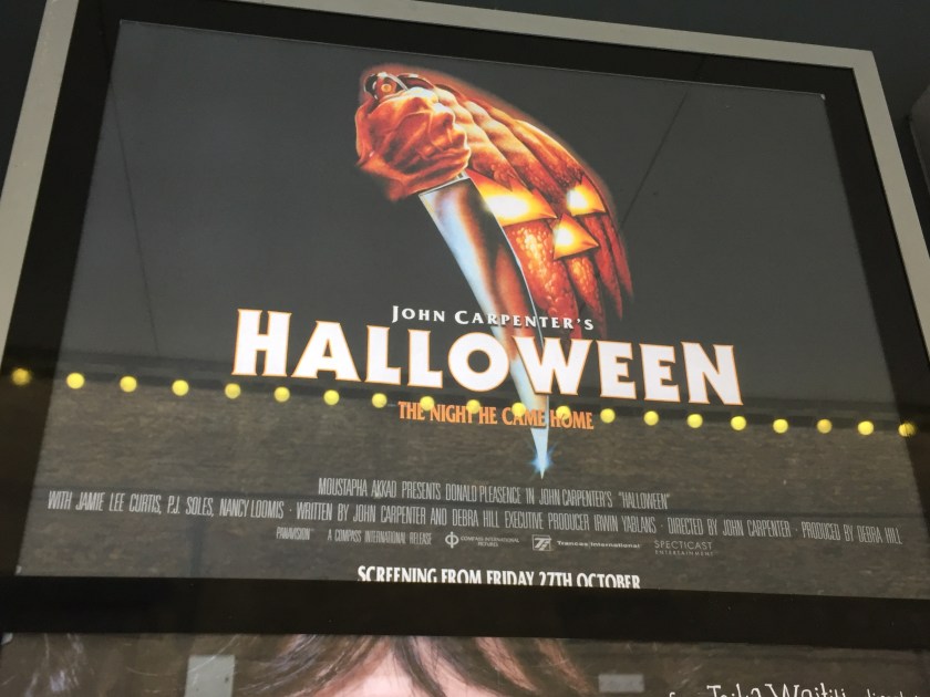 Poster outside cinema for John Carpenter's Halloween. Above the movie title is a large masked face with a hand holding a large knife pointed downwards.