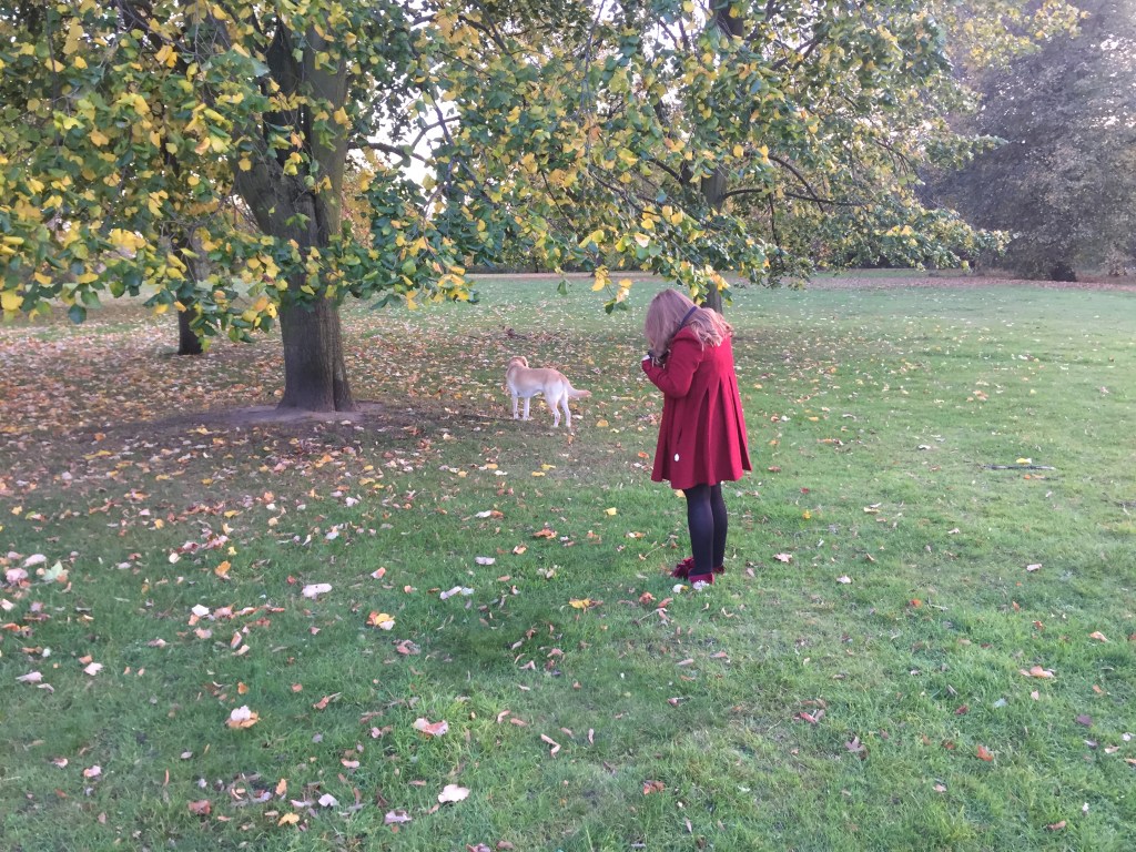 Emily Davison, wearing a red coat and standing beneath a large tree in Hyde Park, with her golden labrador guide dog Unity ahead of her looking around.